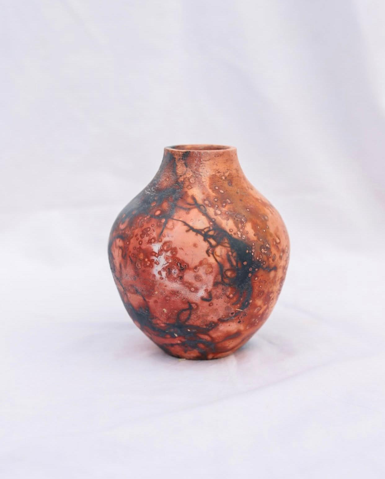 Gerro de ceràmica de línies corbes amb una textura i coloració orgànica molt intensa en tons vermell foc, carabassa i taques negres abstractes, sobre un fons blanc pur.