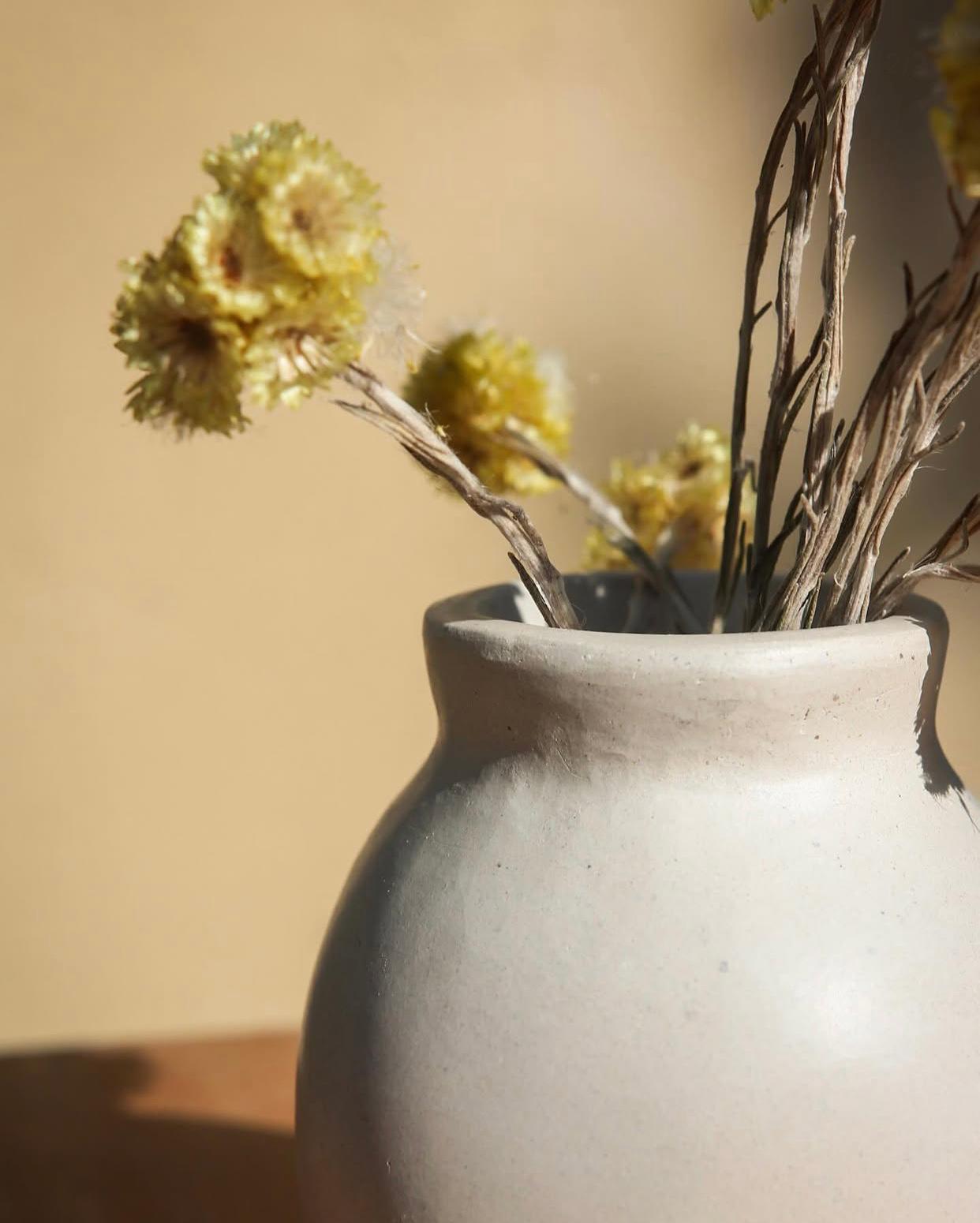 Detall del coll d'un gerro de ceràmica blanca d'acabat mat i rústic, decorat amb flors seques grogues i il·luminat per una llum càlida i suau.