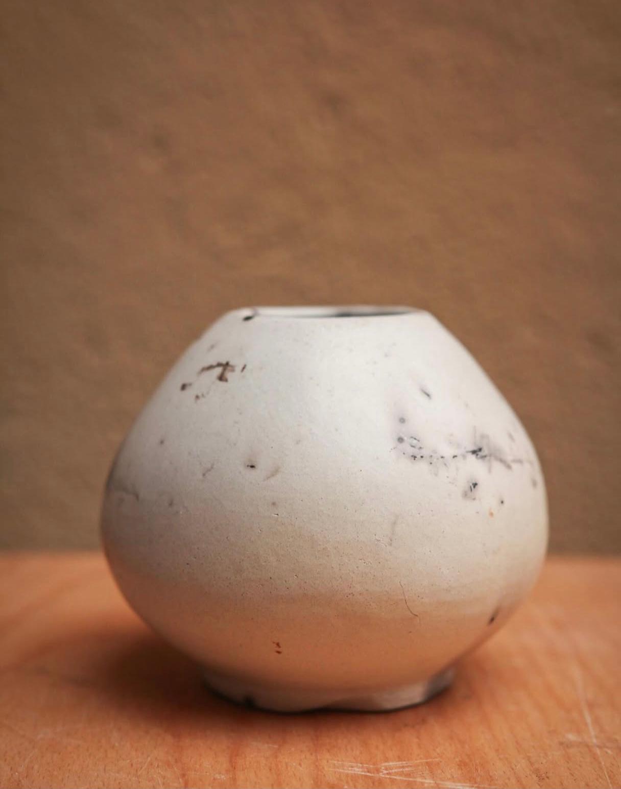 Gerro rodó de ceràmica clara d'estil minimalista i orgànic, amb petites marques grises i negres del procés de cocció, reposant sobre una superfície de fusta.