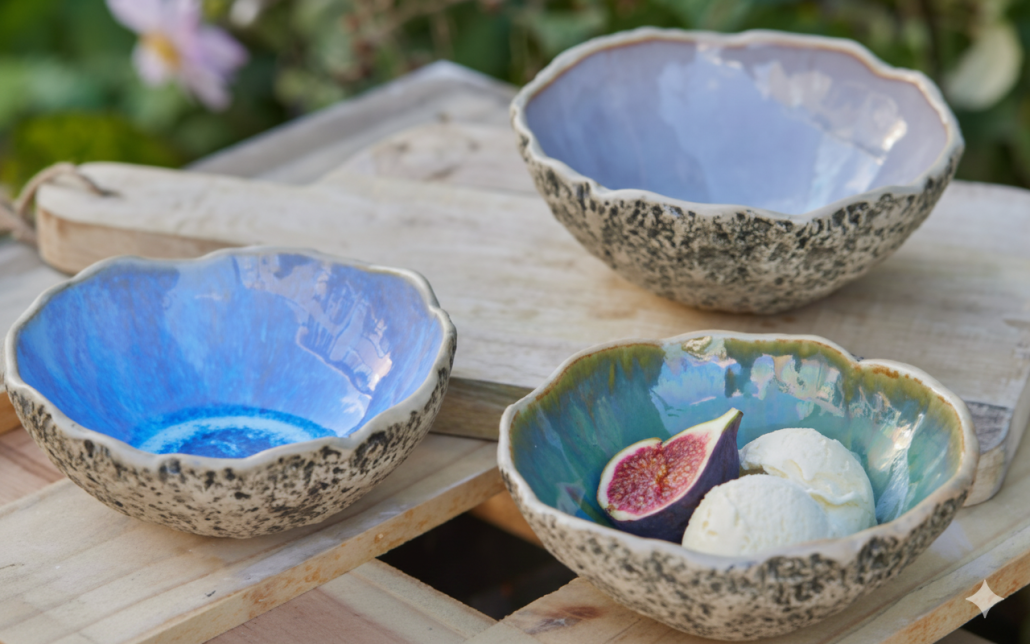 Generated Image April 14, 2026 - 11_39AM Conjunt de tres bols de ceràmica artesanal amb l'exterior texturat i l'interior esmaltat en tons blaus, liles i verds vibrants, presentats amb figues sobre fusta.